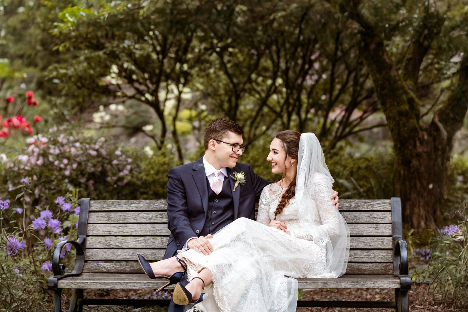 Newlyweds share intimate moment on garden bench surrounded by blooming flowers.