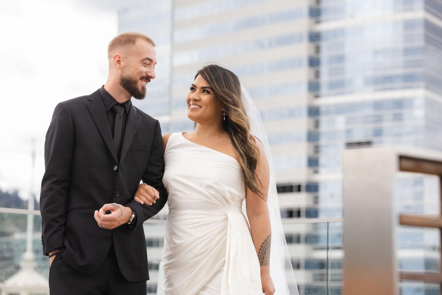 A couple in formal attire poses together on a rooftop with modern city buildings in the background.
