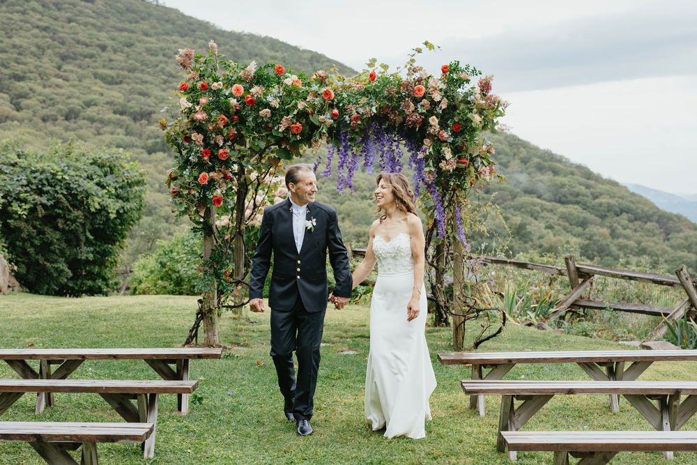 Couple walks along scenic mountain path with wooden benches and flowering bushes during outdoor wedding celebration.