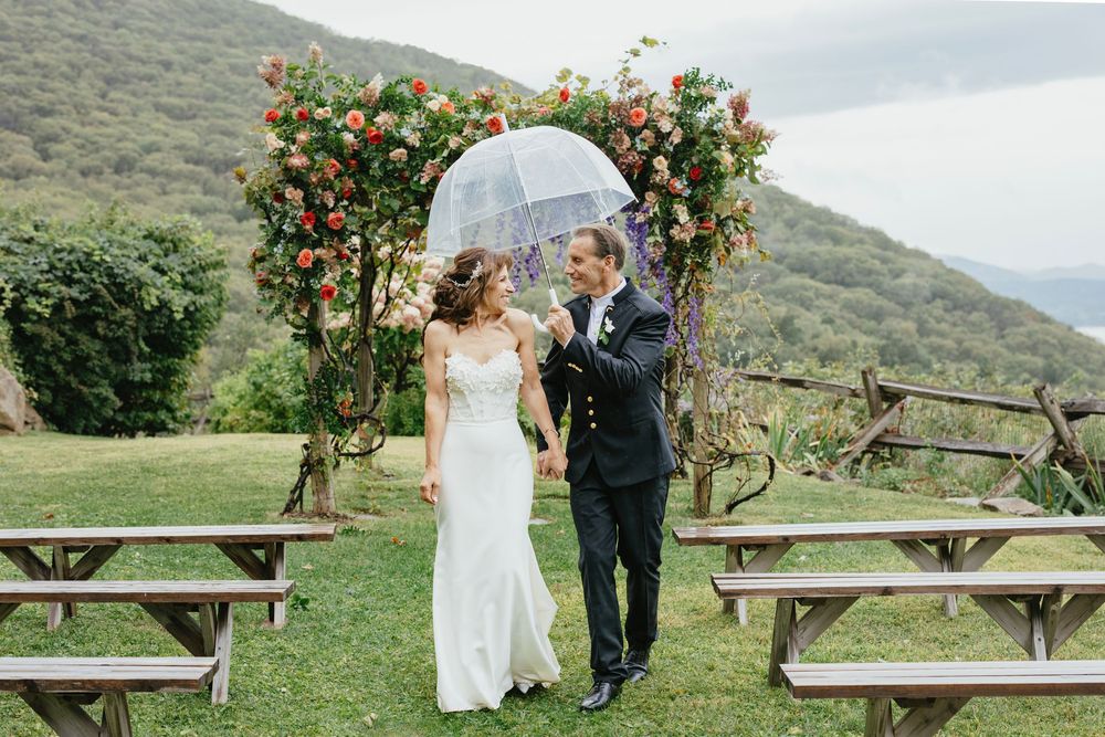 A romantic outdoor wedding ceremony takes place beneath a floral arch on a scenic hillside with picnic tables nearby.