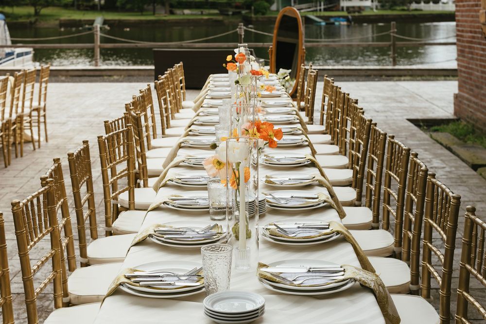 Elegant outdoor wedding reception table setup with gold chairs and peach floral centerpieces beside a lake.