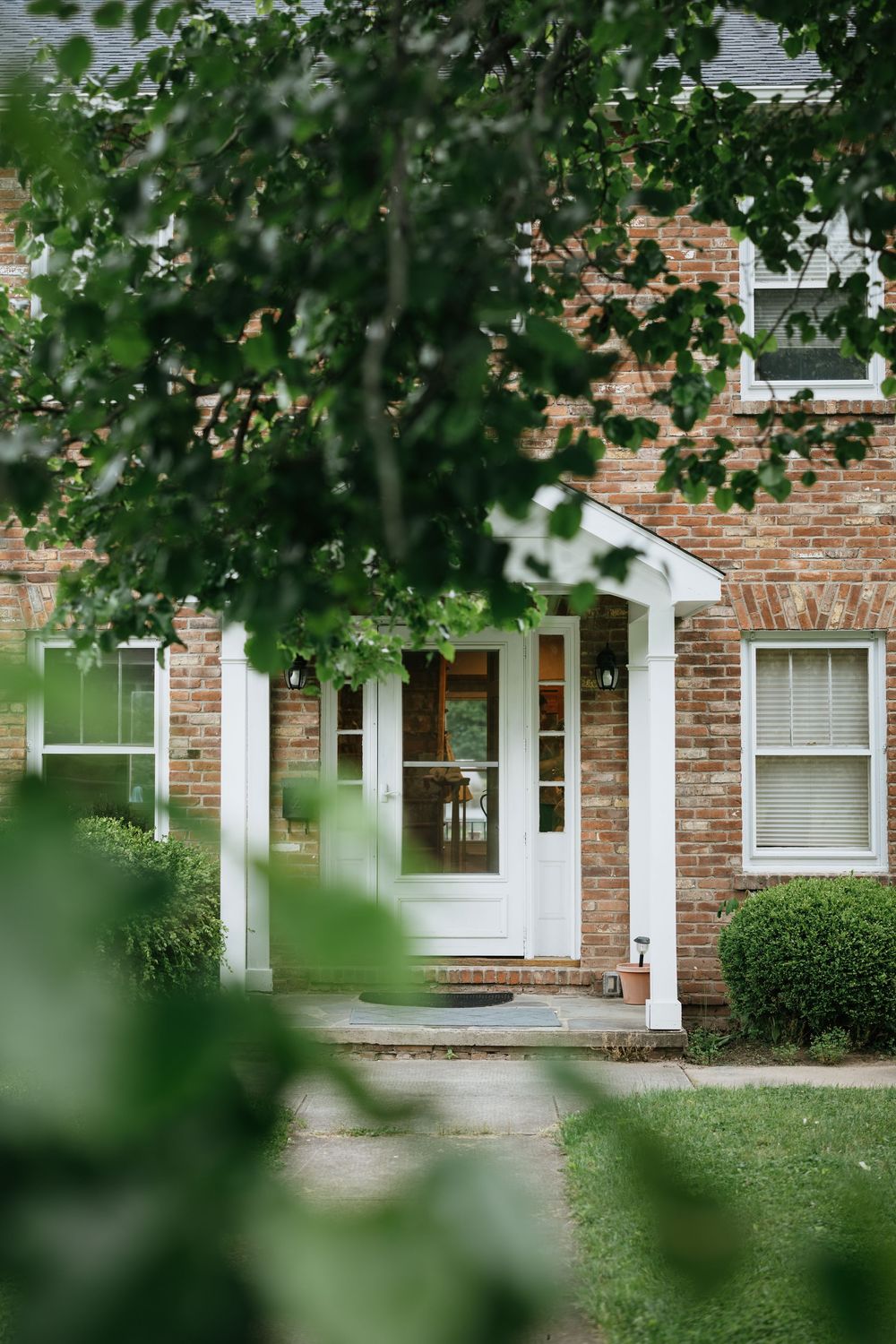 Partial view of a brick residential building with white trim and windows seen through green foliage.