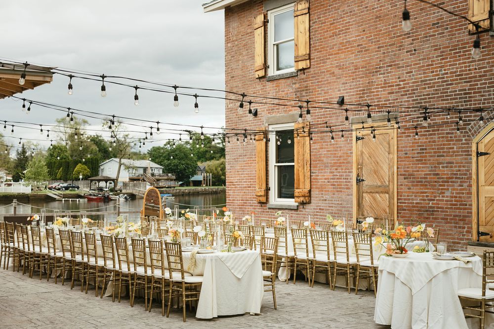 String lights illuminate a rustic outdoor reception setup with wooden chairs and white tablecloths against a brick building.