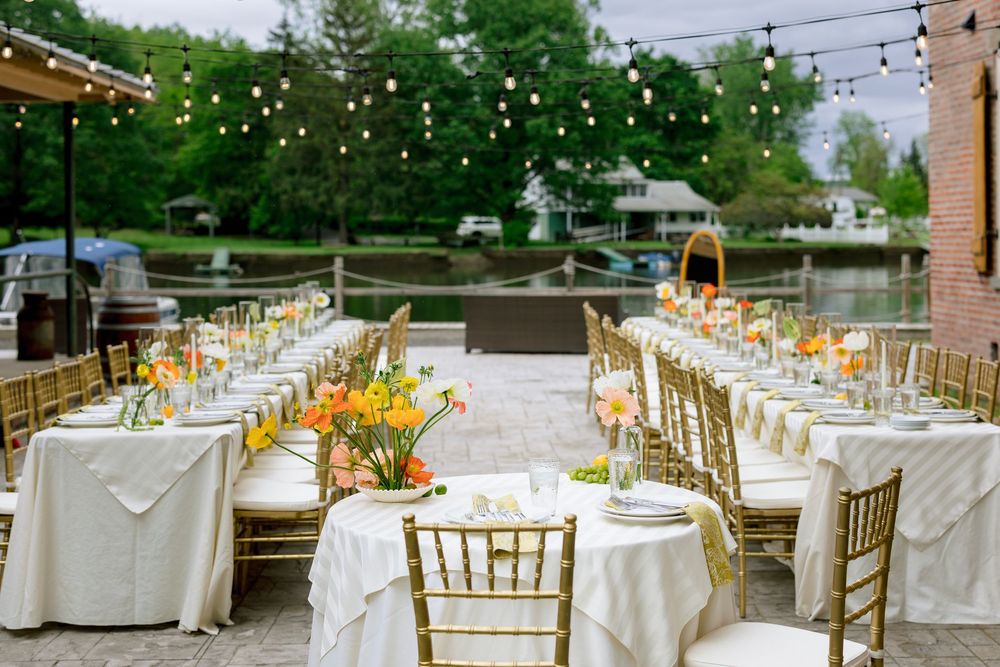 Elegant outdoor wedding reception setup with long tables, gold chairs and string lights under a covered pavilion.