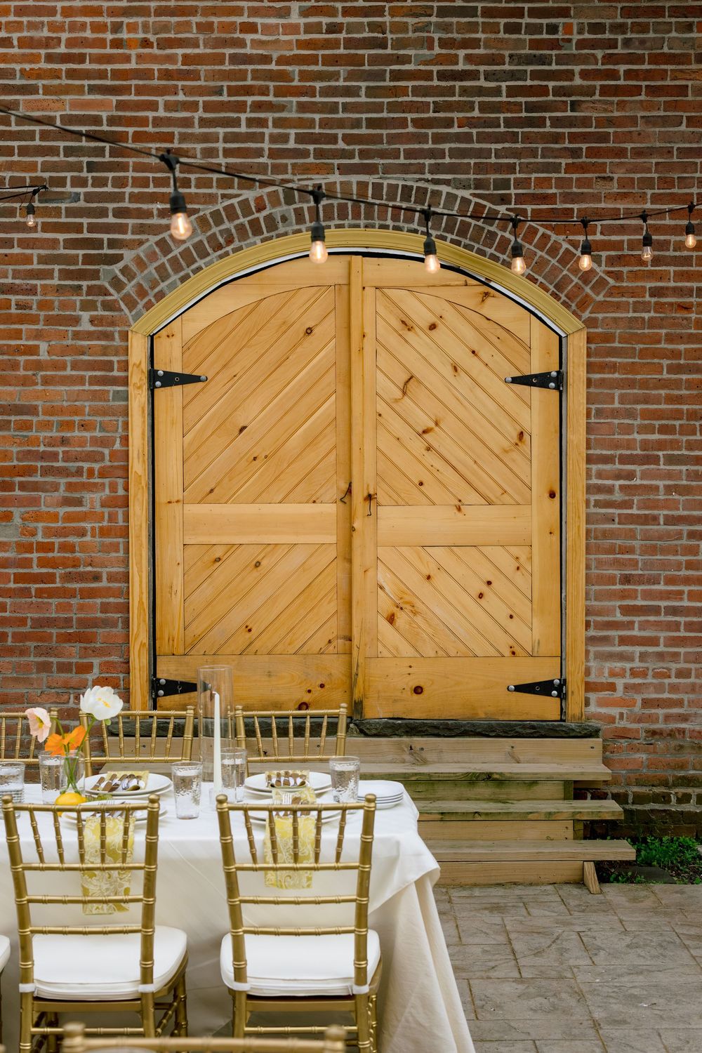 Large wooden barn doors with chevron patterns mounted on a rustic brick wall with elegant dining chairs arranged outside.