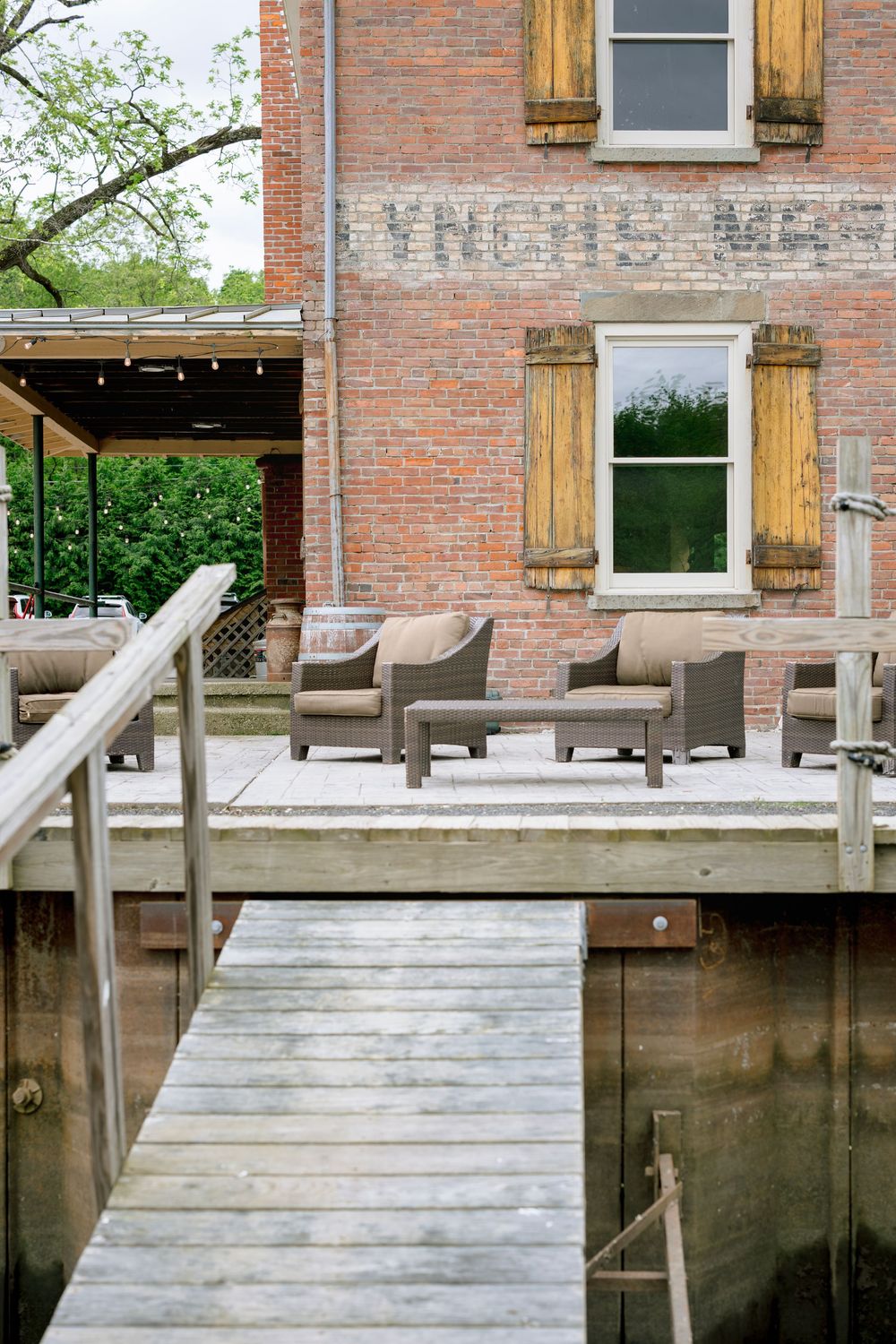 A rustic wooden boat dock with steps leads up to a brick building with string lights overhead and outdoor seating.