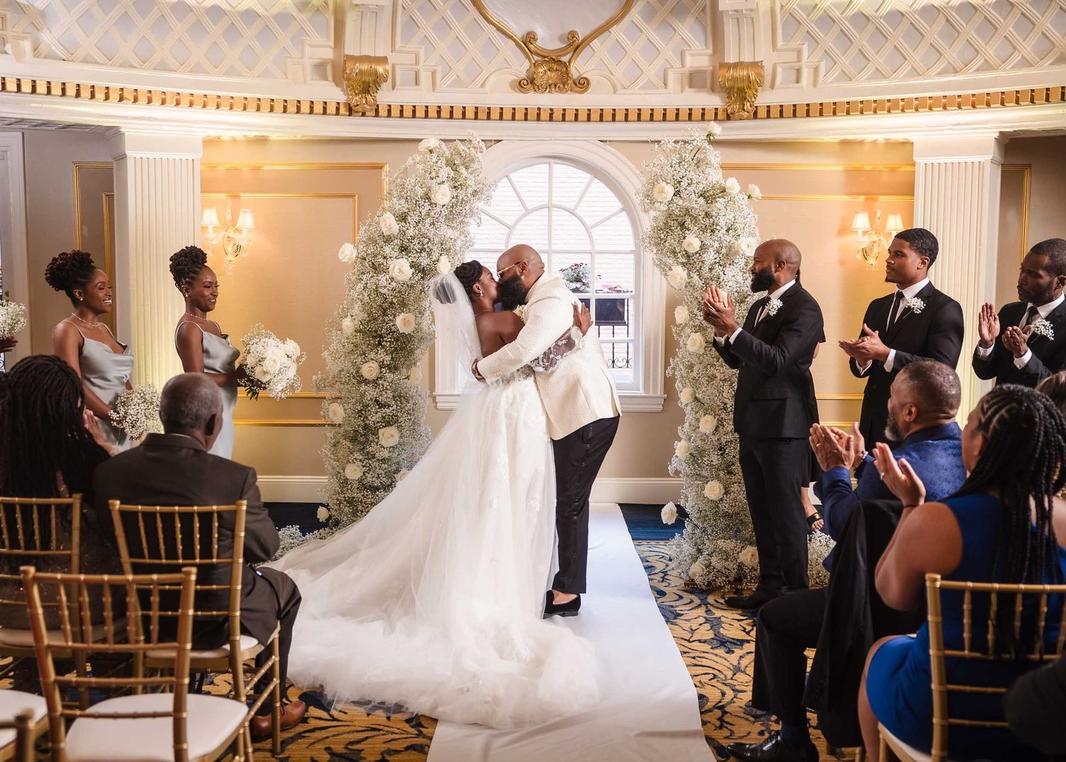 Bride and groom first kiss under floral arch in the Dome Room at Lenox Hotel Boston with guests applauding