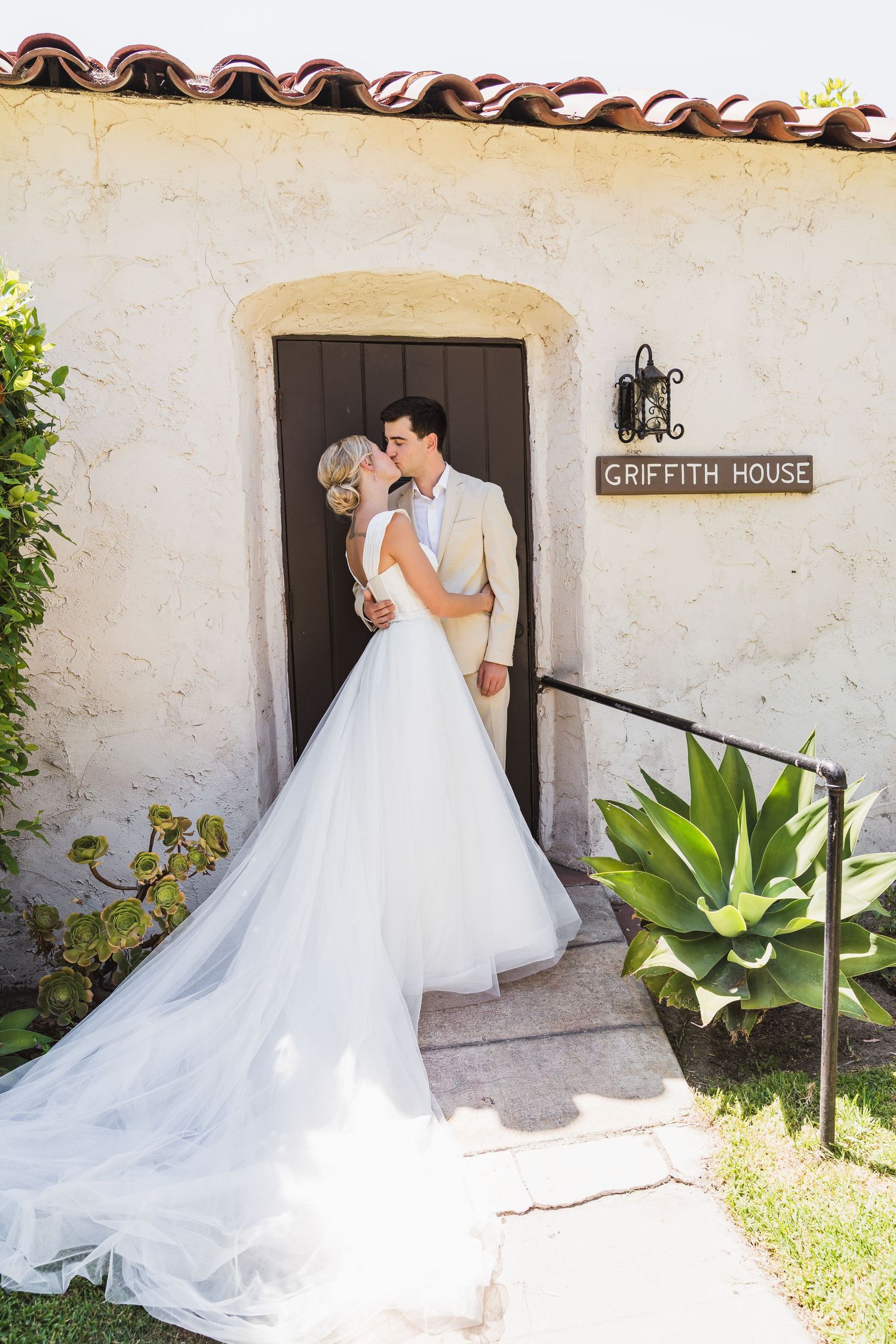A romantic wedding couple shares a tender moment outside the historic Griffith House with lush agave plants in the garden.