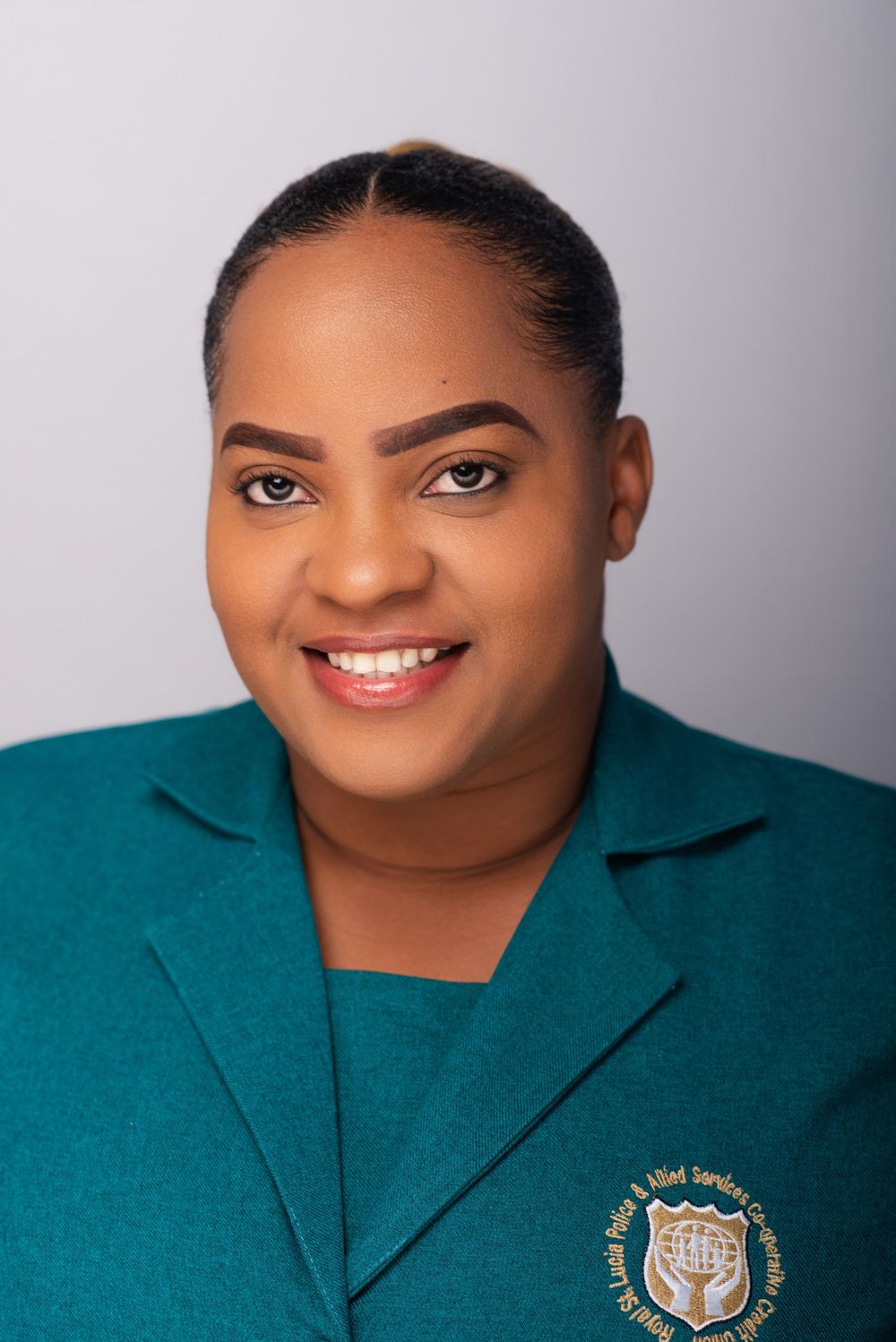 Professional headshot of someone wearing a teal blazer with an embroidered logo against a gray background.