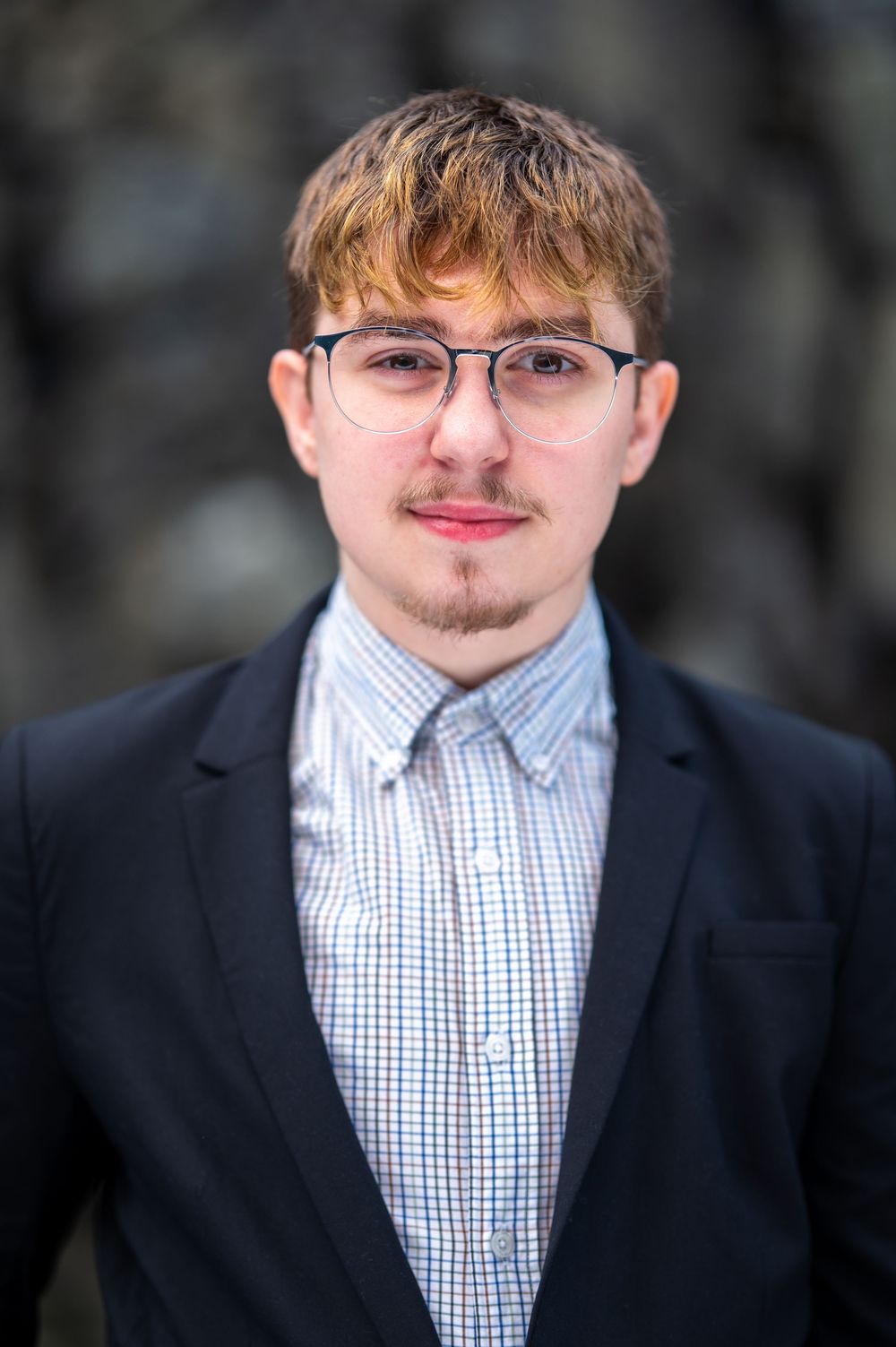 Professional headshot of a person wearing glasses, a checkered button-down shirt and dark blazer against a blurred background.