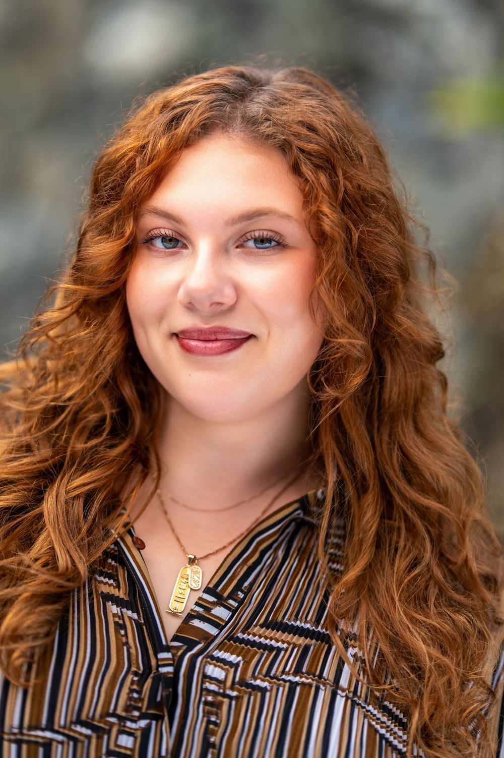 A person with long curly red hair wearing a black and white striped shirt smiles at the camera.