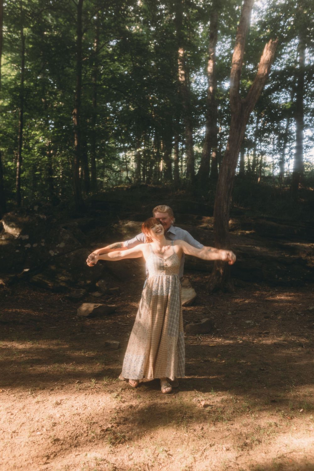 Couple dancing together in sunlit forest clearing, woman in white lace dress with arms outstretched.