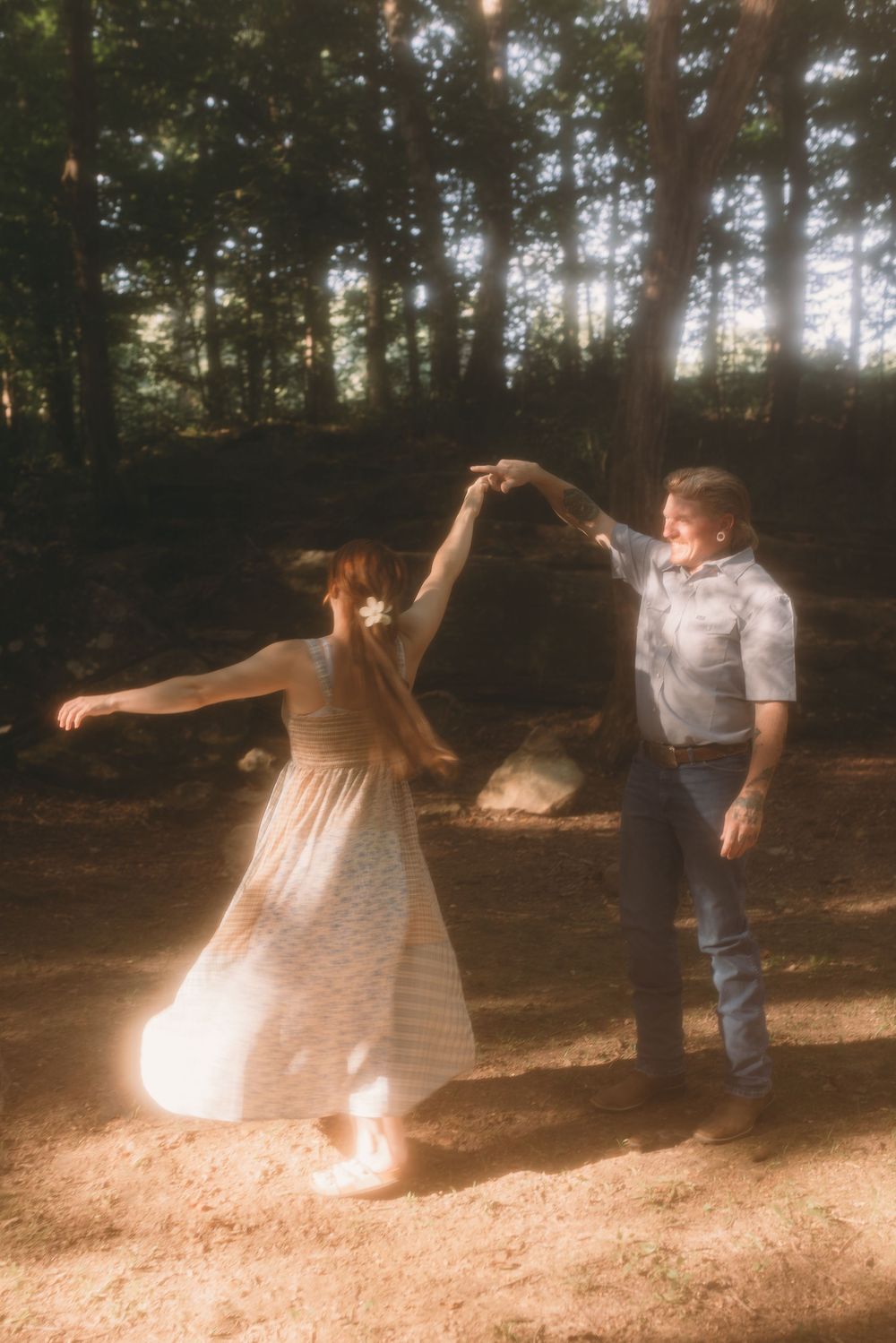 Couple dancing together in a sun-dappled forest, woman in flowing white dress being twirled.
