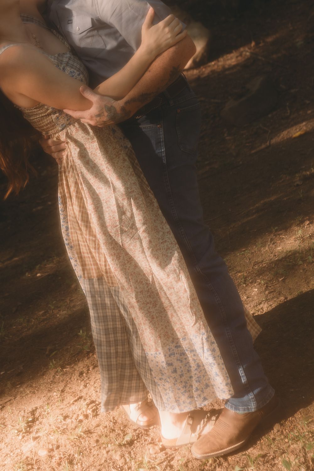 Couple embracing outdoors, woman in floral maxi skirt and man in dark jeans, standing on sunlit ground.