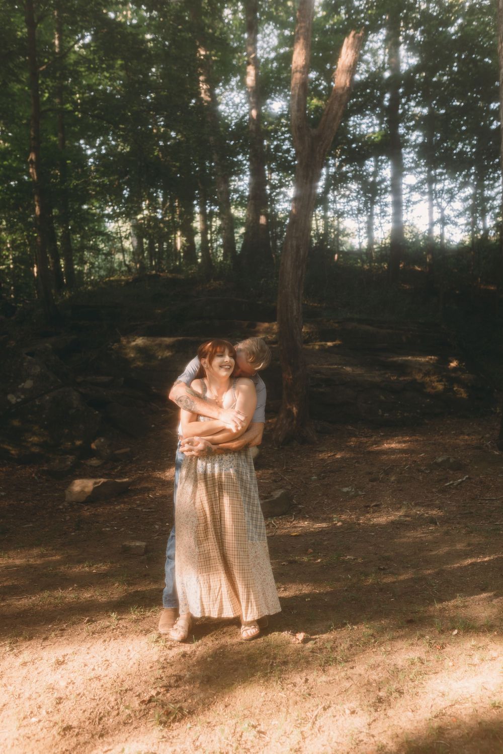 Couple embracing and kissing in a sunlit forest clearing surrounded by tall trees.