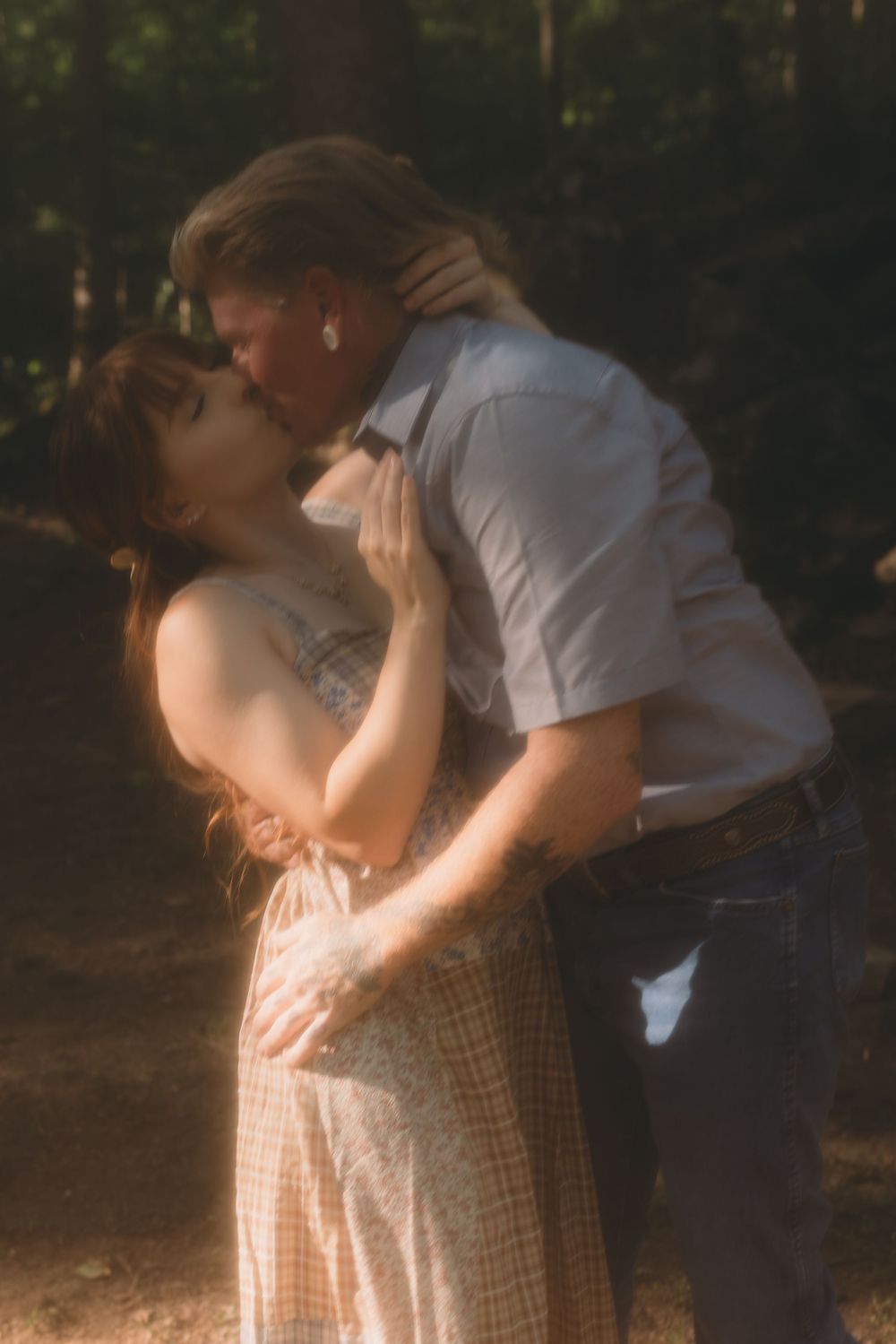 A couple shares a romantic kiss in a sun-dappled forest, the woman in a floral dress embracing the man tenderly.