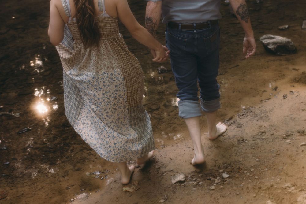 Barefoot couple holding hands walking on rocky path near water, woman in floral dress and man in jeans.
