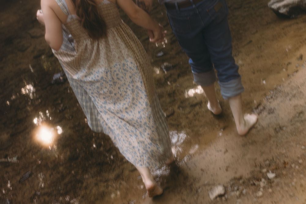 Couple walking barefoot on wet ground, woman in floral dress and man in jeans, with sparklers at night.