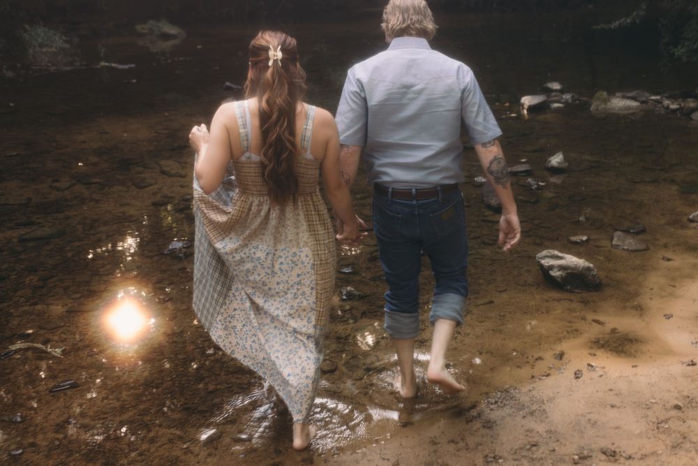 Couple walking barefoot hand in hand through a muddy creek bed surrounded by rocks and nature.