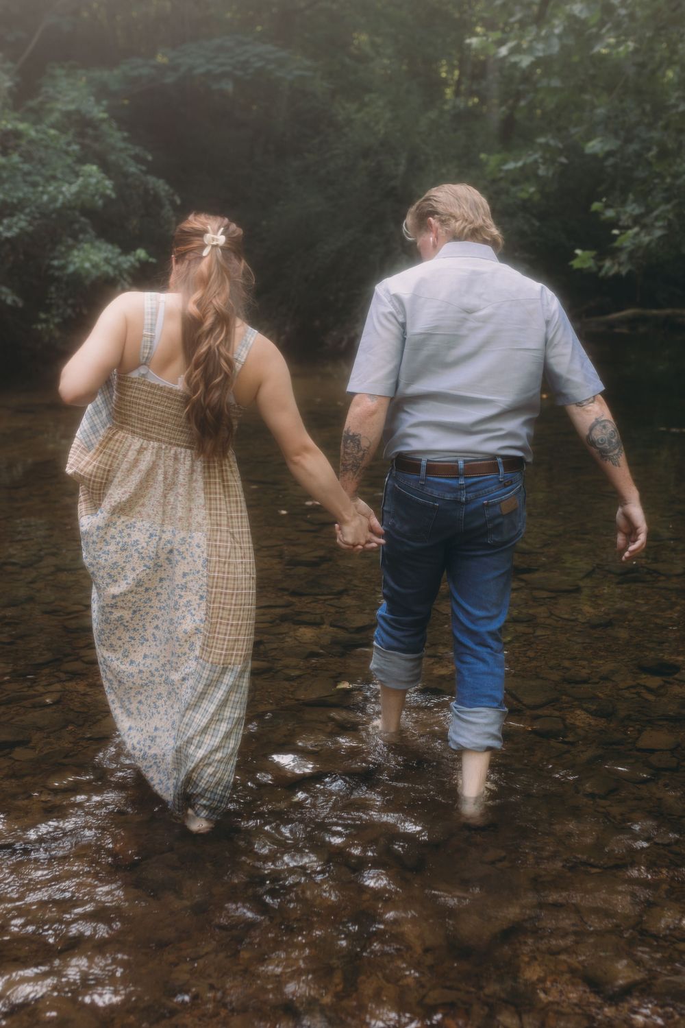 A couple holds hands while wading barefoot through a shallow forest creek from behind.