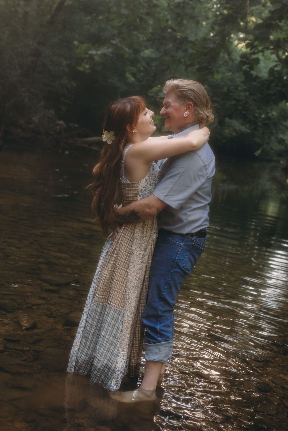 Couple embracing romantically while standing in a shallow forest creek, woman in floral dress with long red hair.