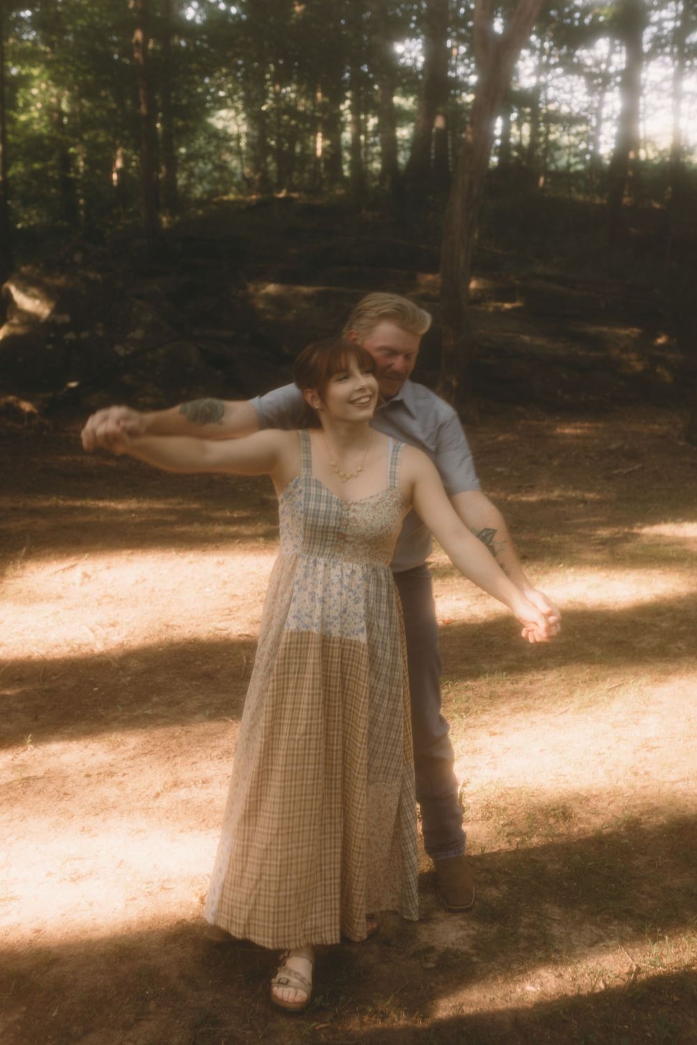 A smiling woman in a plaid midi dress dances with her partner in a sun-dappled woodland setting.