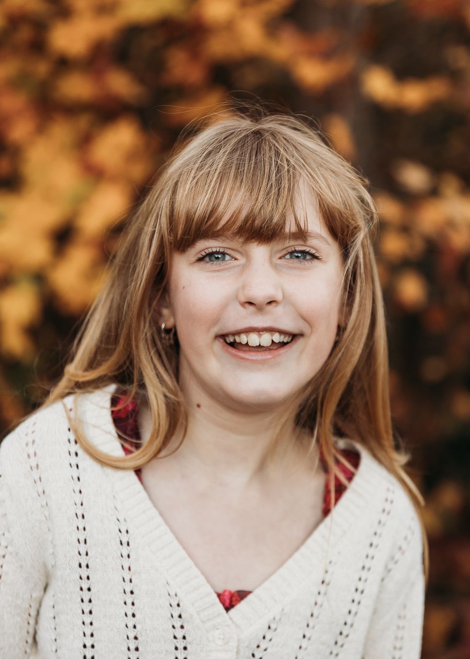 A smiling person in a white knit sweater poses outdoors against warm autumn foliage in the background.