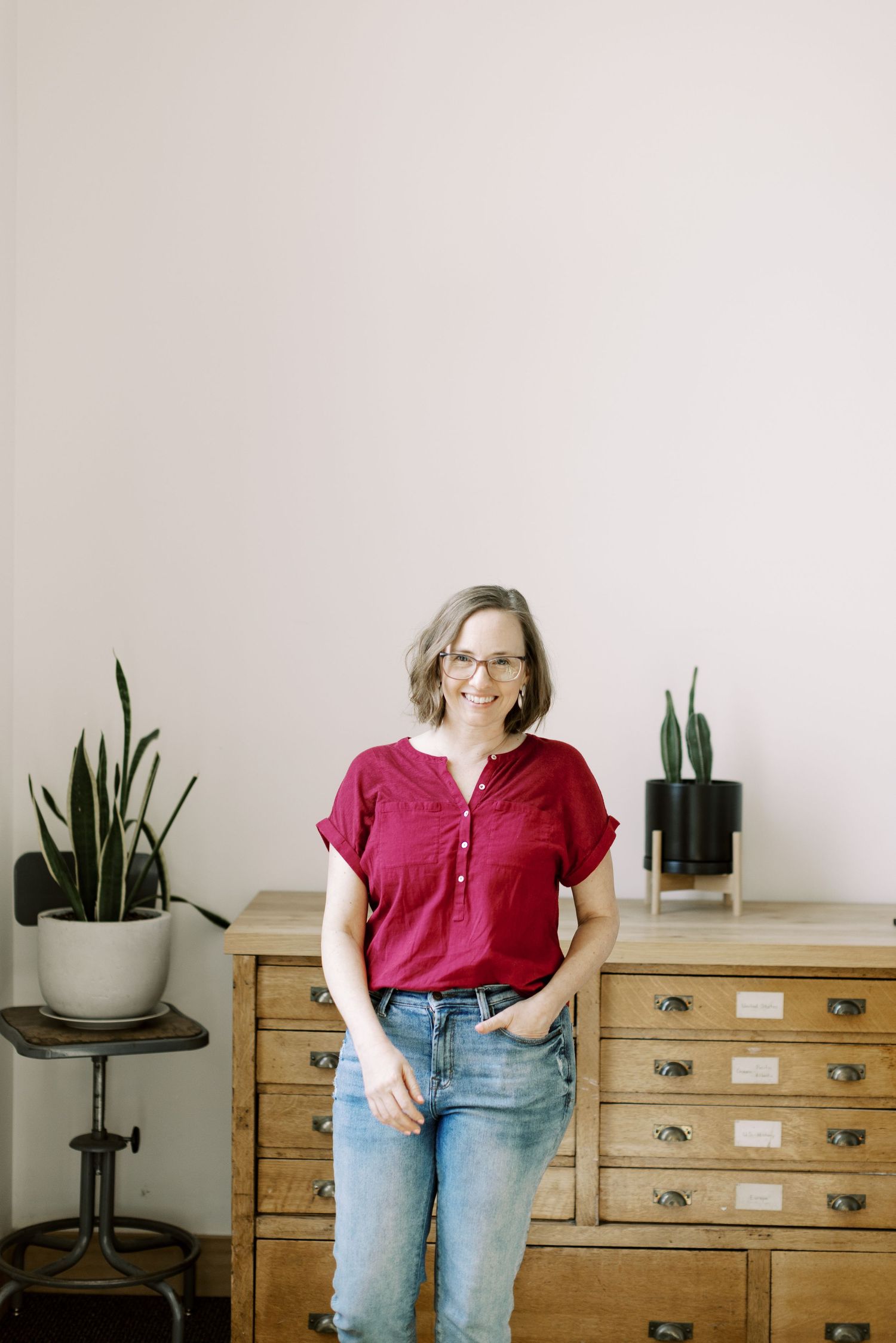 Woman in red top and jeans stands casually by wooden dresser with houseplants.