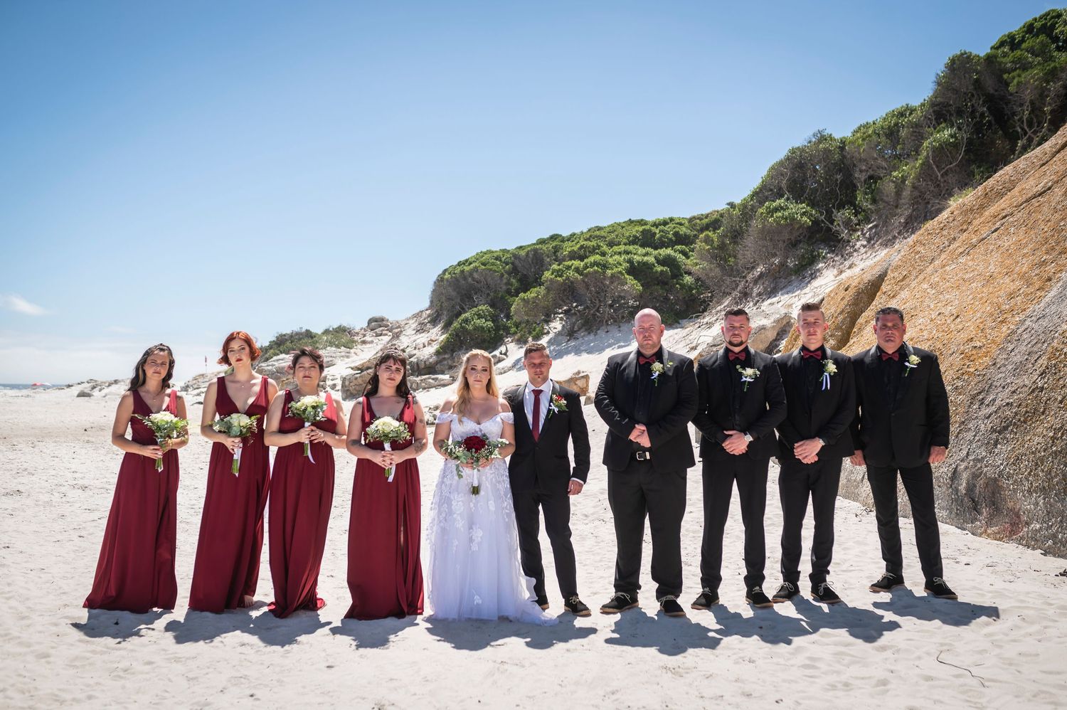 Wedding party posing on a sandy beach, bridesmaids in red gowns and groomsmen in black suits with bride and groom.