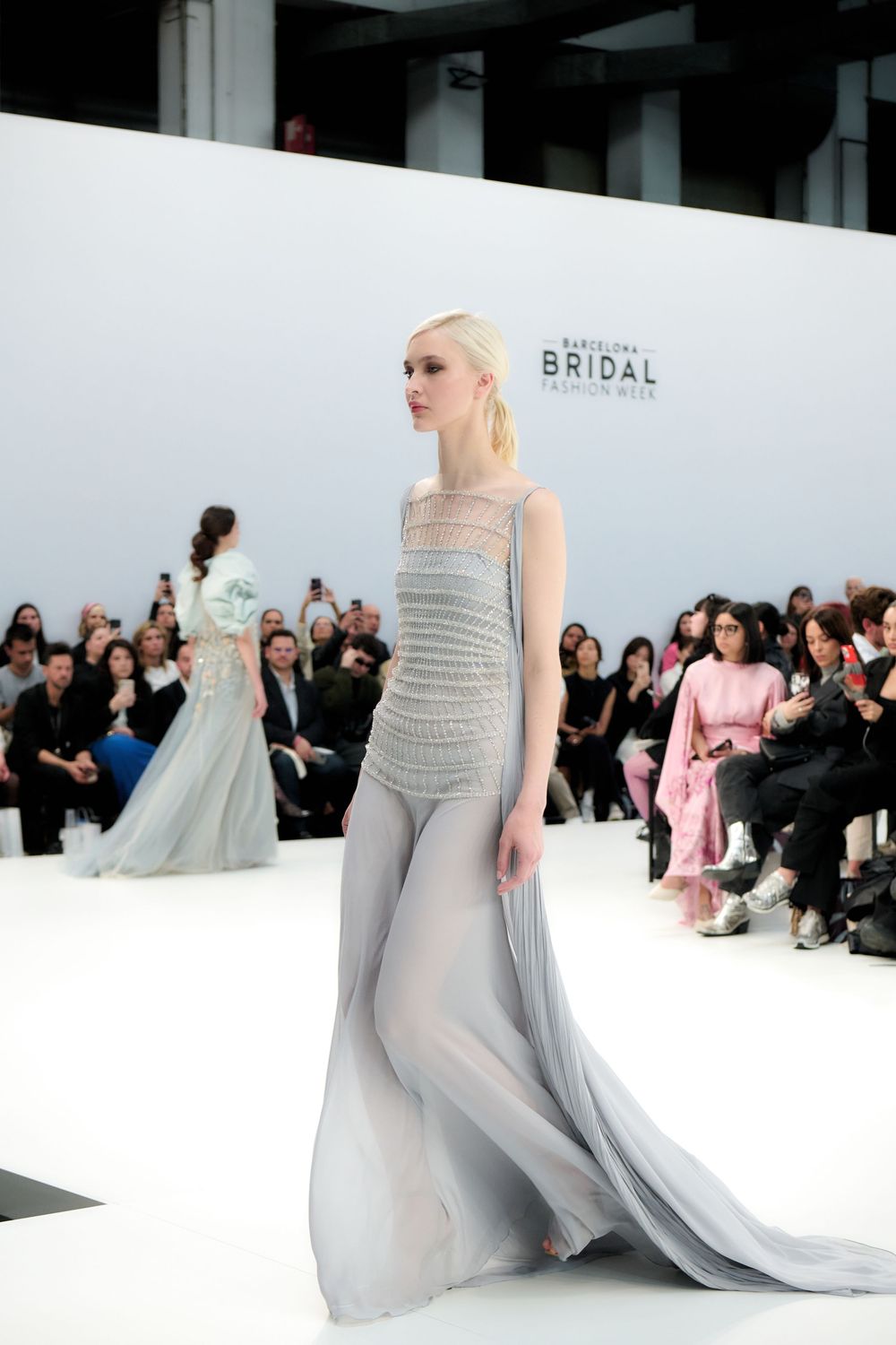 Ethereal grey bridal gown showcased at fashion week with crowd in background.