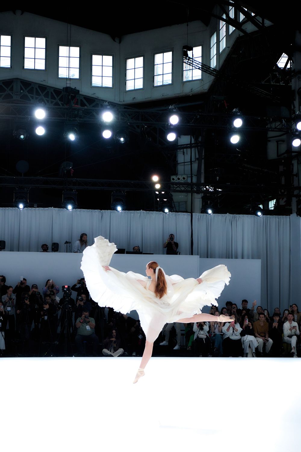 Bridal fashion show featuring flowing white gowns on a stark white runway with dramatic overhead lighting.