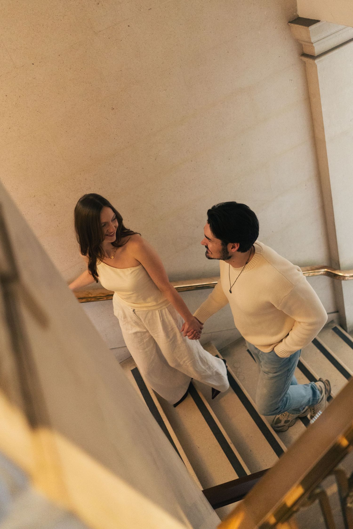 Couple holding hands on museum stairs, sharing a smile in a warm golden-lit stairwell.