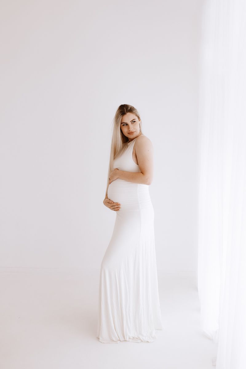 Elegant maternity portrait in white dress against minimalist background.