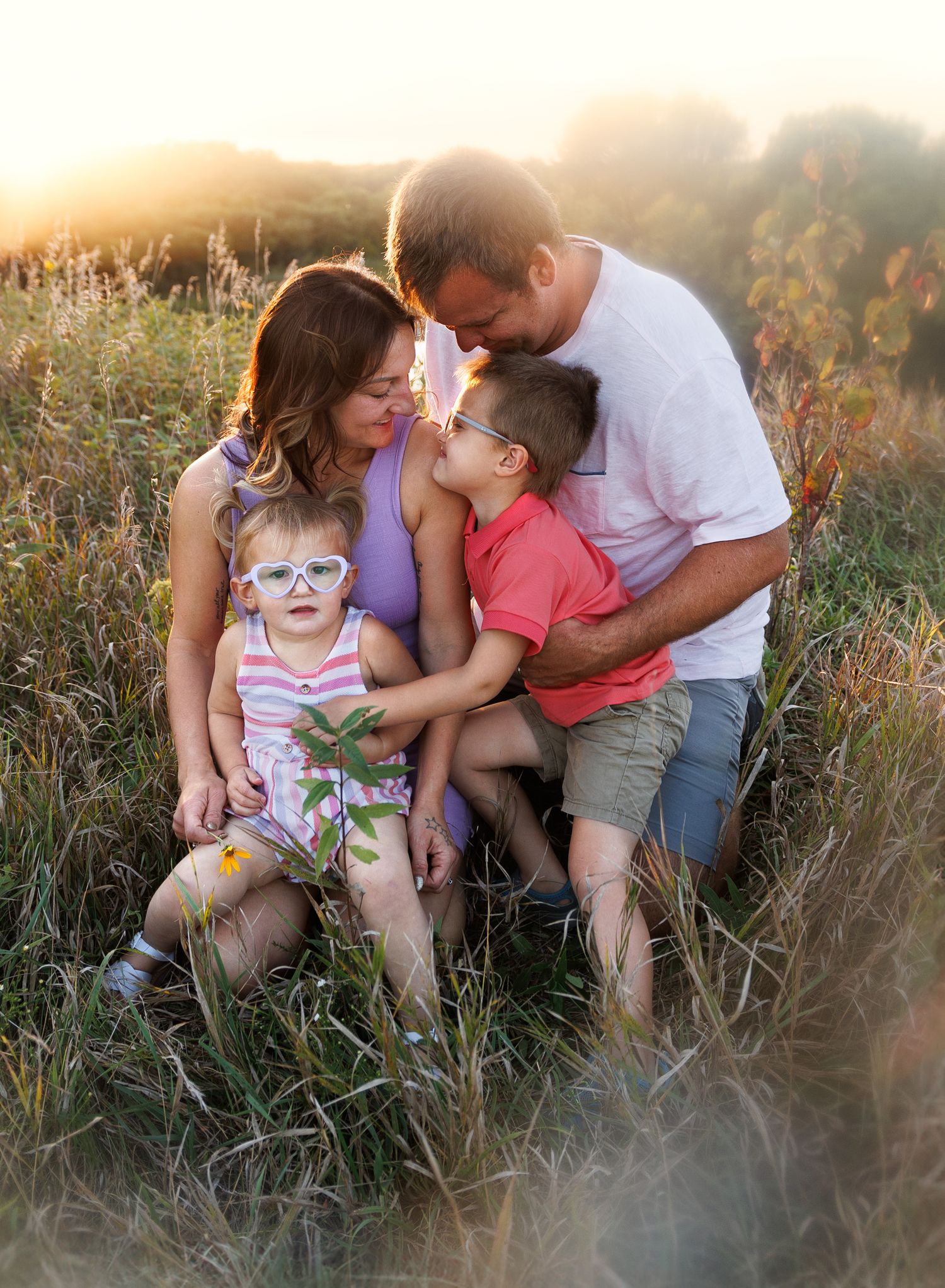 A family sits together in a sunlit meadow during golden hour as tall grass sways around them in the warm summer breeze.