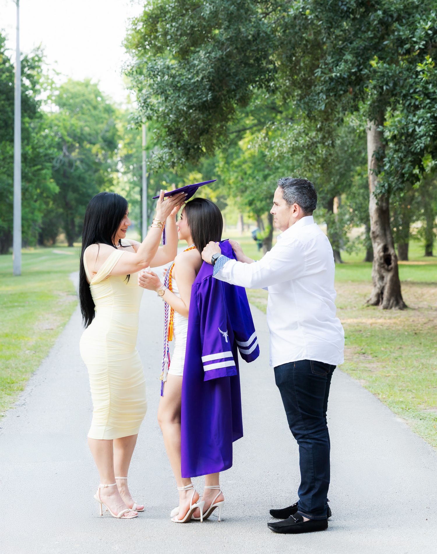 A family celebrates graduation on a tree-lined path in a park with a graduate in purple regalia getting ready.