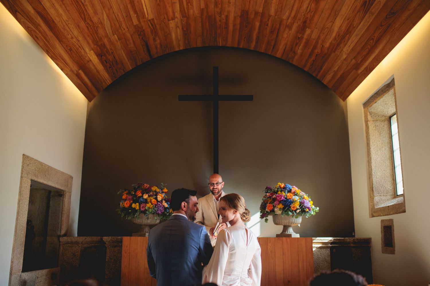 A small wooden chapel interior with vaulted ceilings, cross mounted on wall, and colorful floral decorations.
