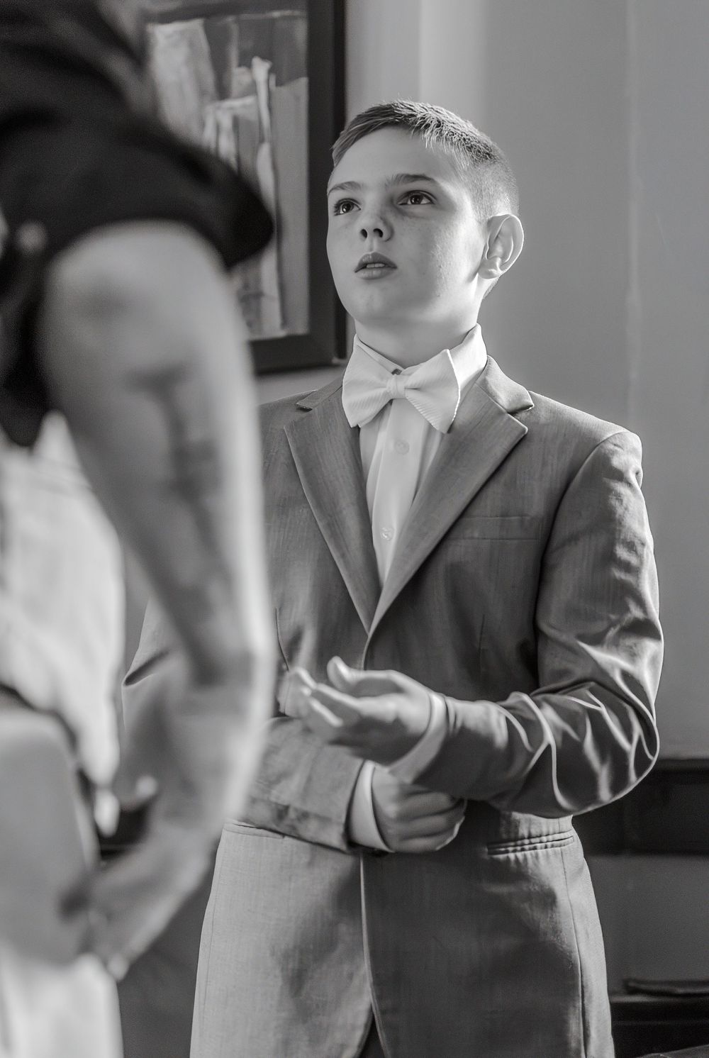 Black and white photo of a young boy in a suit and bow tie looking up attentively at a wedding ceremony.
