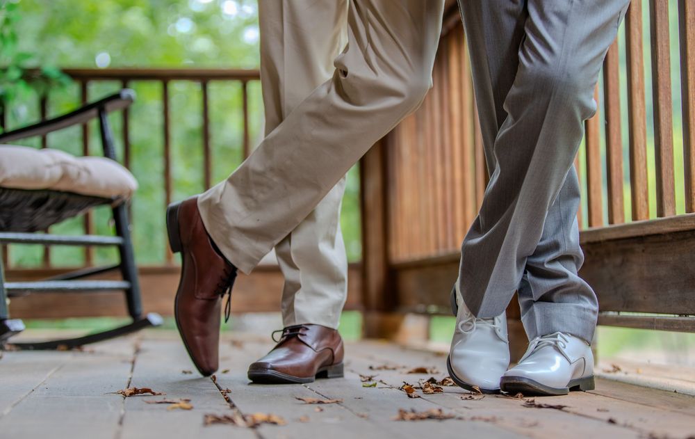 Two men in dress shoes stand on a wooden deck, one wearing brown oxfords with khakis, the other in white shoes with gray pants.