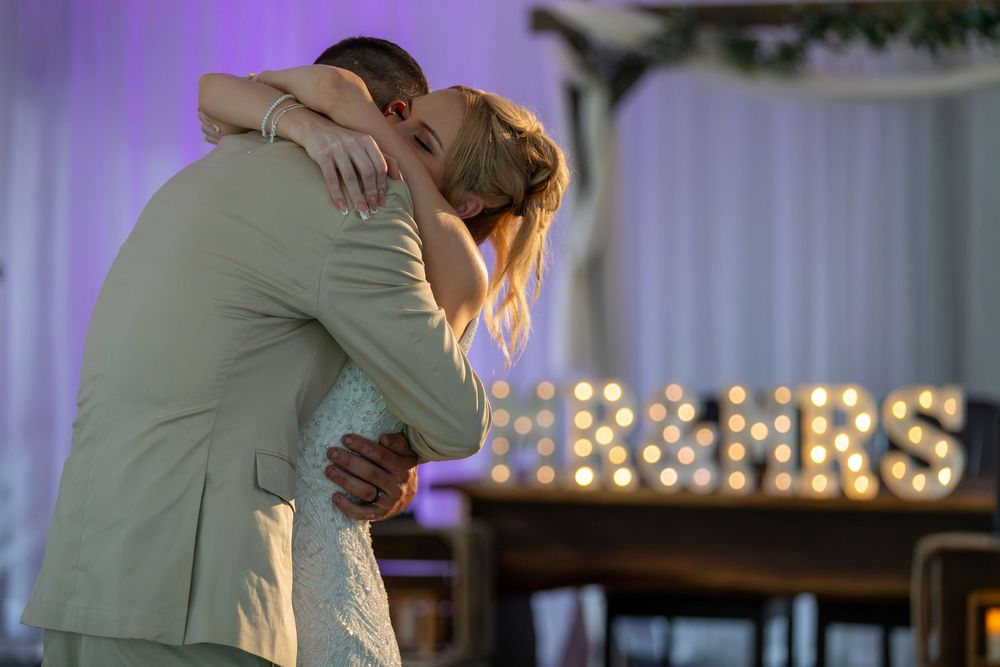 Newlywed couple sharing first dance embrace in front of illuminated MR & MRS marquee letters at wedding reception.