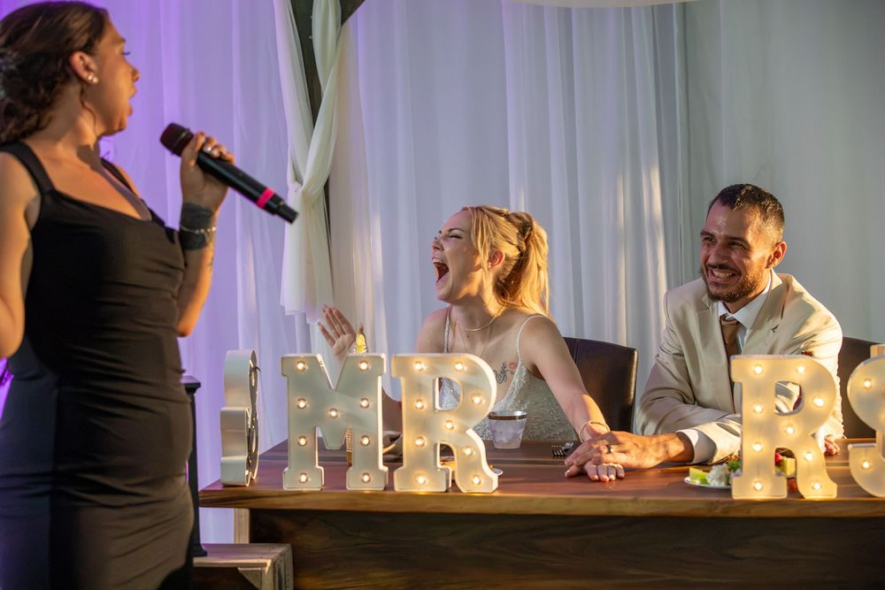 Bride and groom laugh at wedding reception table as woman in black dress speaks into microphone near MR & MRS letter lights.