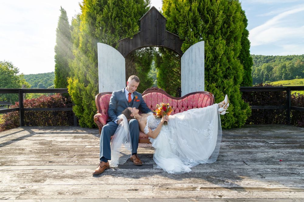 Bride and groom laughing on vintage pink sofa outdoors at rustic wedding venue with lush greenery backdrop.
