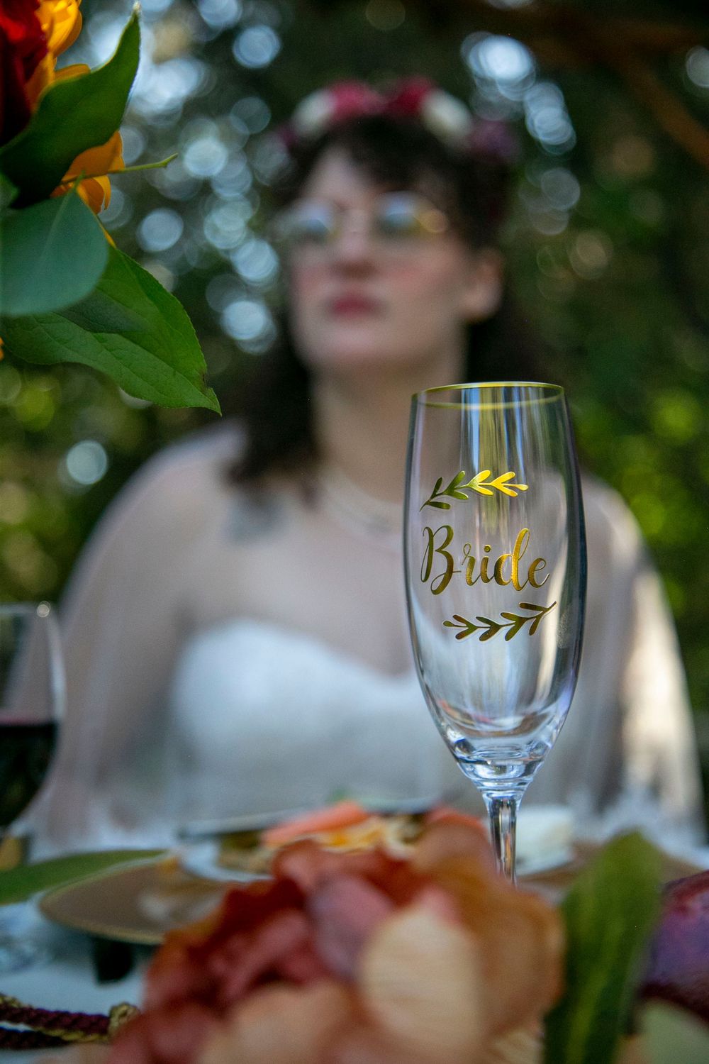 A crystal champagne flute engraved with Bride in gold sits on a table with a blurred bride in the background.