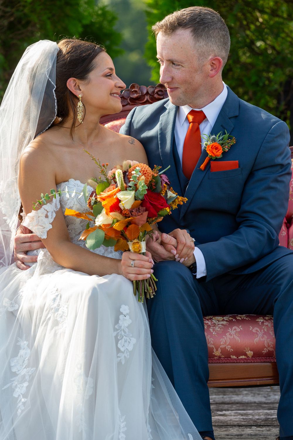 Bride and groom in blue suit with orange tie share a loving gaze, holding a vibrant autumn bouquet outdoors.