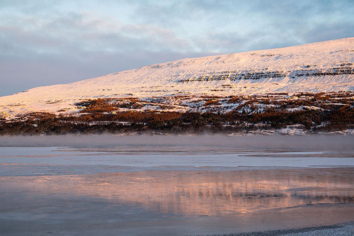 Snow-covered mountains reflect in calm lake waters during sunset creating a peaceful winter landscape.