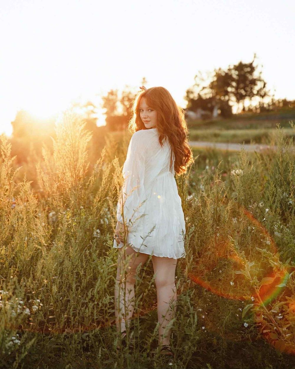 A high school senior girl in a white dress twirls through a sunlit meadow at golden hour as tall grasses sway in the breeze.