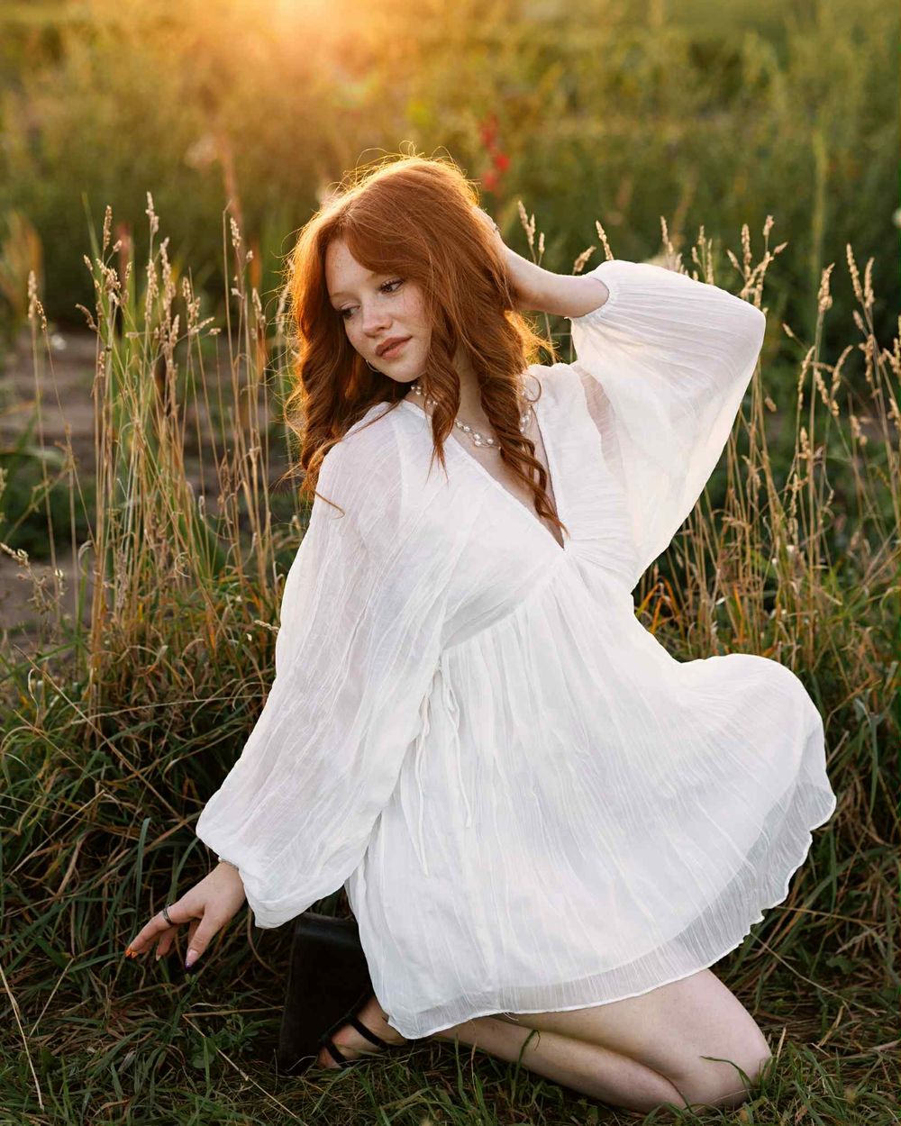 A dreamy sunset portrait in a field of tall grass with a flowing white dress and red hair catching the golden light.