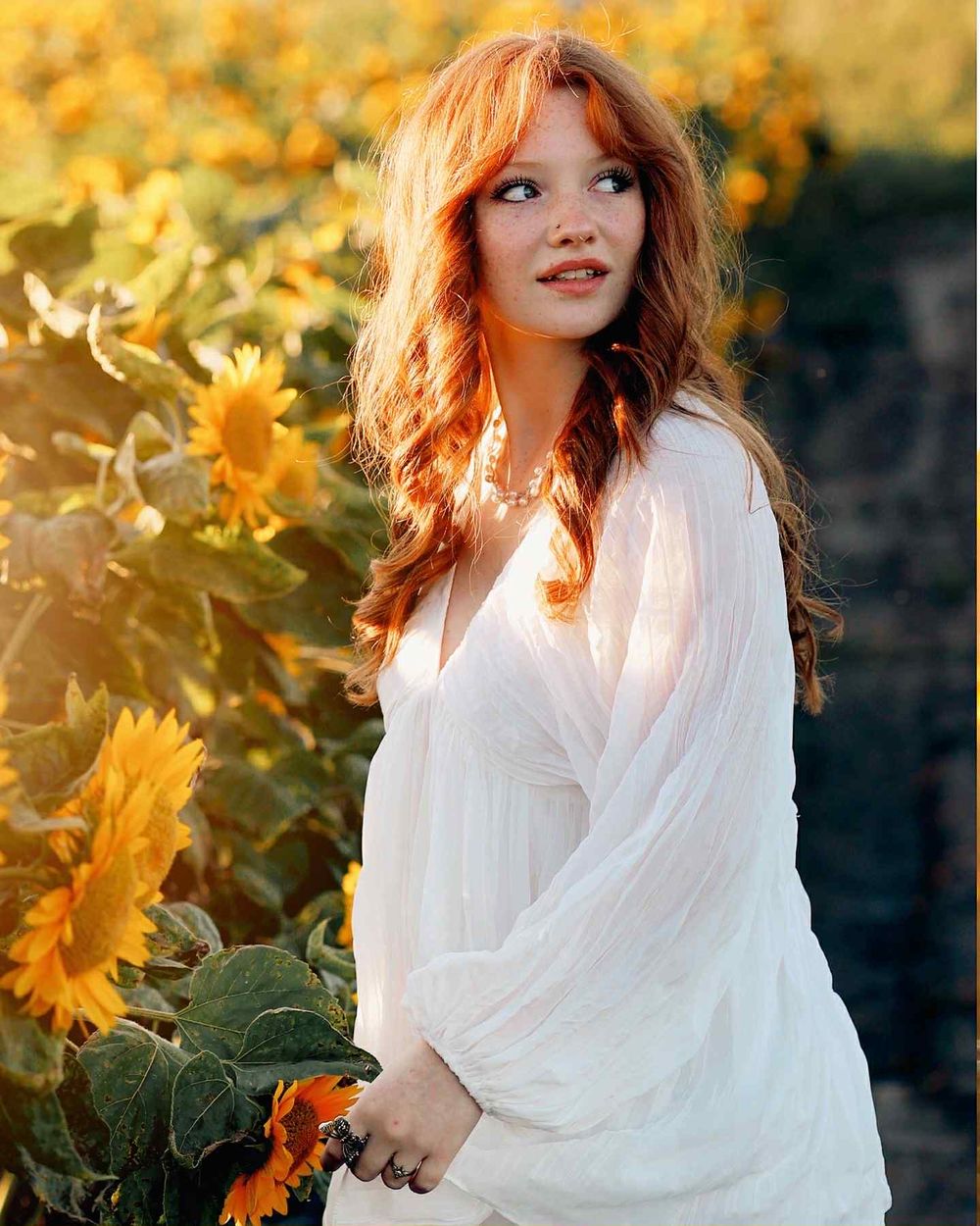 A high school senior in a flowing white dress poses in a sunflower field at golden hour with warm autumn lighting at Bill's Farmers Market