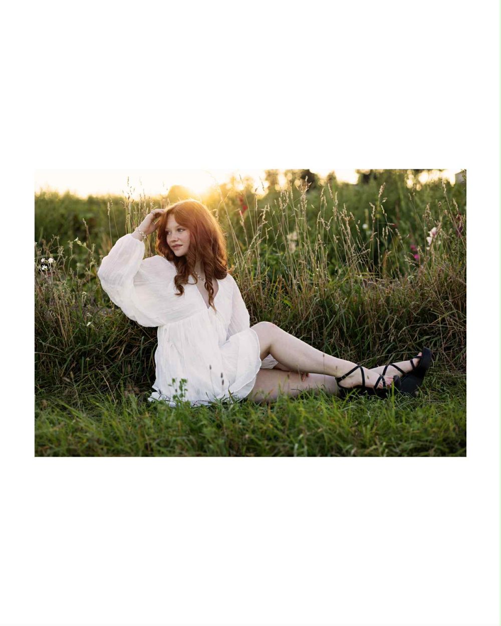 Twelfth grader in a white dress lying gracefully in a sunlit meadow with tall grass at sunset.