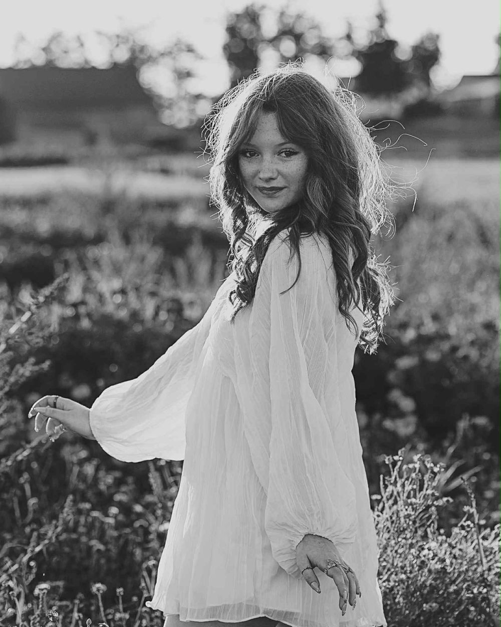 A high school senior in a flowing white dress twirls gracefully in a sunlit meadow with wildflowers in black and white in Petoskey