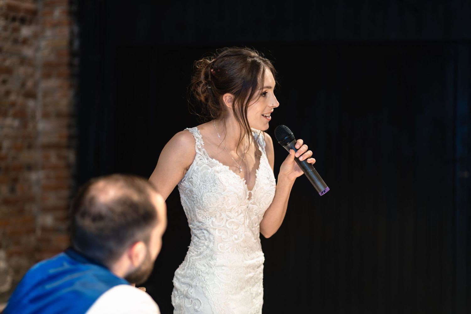 A bride in a white lace dress sings into a microphone during a wedding reception.